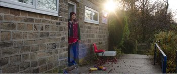 Movie still from “Adult Life Skills” (2016), directed by Rachel Tunnard – A woman leaning against a brick wall near a red chair; Wide shot, High angle