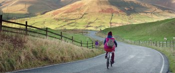 Movie still from “Adult Life Skills” (2016), directed by Rachel Tunnard – A person riding a bike down a road near a grassy hill; Extreme Wide shot, High angle