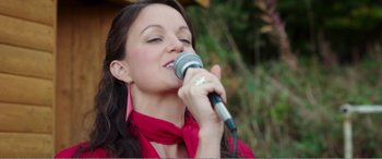 Movie still from “Adult Life Skills” (2016), directed by Rachel Tunnard – A woman in a red shirt is singing into a microphone; Close Up shot, Low angle