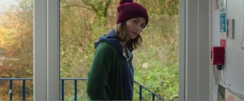 Movie still from “Adult Life Skills” (2016), directed by Rachel Tunnard – A woman wearing a red hat standing next to a blue railing; Medium shot, Over the shoulder angle