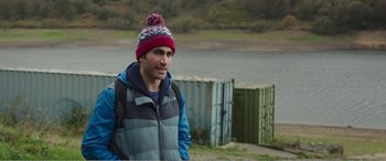 Movie still from “Adult Life Skills” (2016), directed by Rachel Tunnard – A man wearing a jacket and a hat standing next to a body of water; Medium shot, Over the shoulder angle