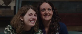 Movie still from “Adult Life Skills” (2016), directed by Rachel Tunnard – Two women smile for the camera while posing for a picture; Close Up shot, Over the shoulder angle