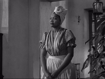 Movie still from “Affair in Trinidad” (1952), directed by Vincent Sherman – A woman wearing a headscarf and apron standing in front of a wall; Medium shot, Low angle