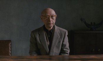 Movie still from “After Life” (1998), directed by Kore-eda Hirokazu – An older man sitting at a table in front of a wall; Medium shot, Low angle