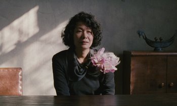 Movie still from “After Life” (1998), directed by Kore-eda Hirokazu – A woman sitting at a table holding a pink flower; Close Up shot, Low angle