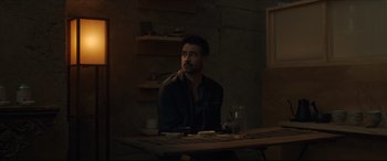 Movie still from “After Yang” (2021), directed by Kogonada – A man sitting at a wooden table in a dark room; Medium shot, Over the shoulder angle