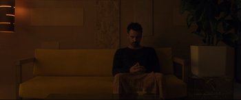 Movie still from “After Yang” (2021), directed by Kogonada – A man sitting on top of a couch in a dark room; Medium shot, Low angle