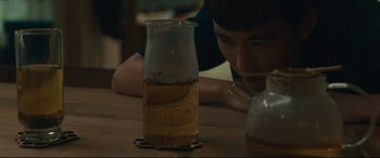 Movie still from “After Yang” (2021), directed by Kogonada – A person sitting at a table with a jar of liquid; Close Up shot, High angle
