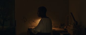 Movie still from “After Yang” (2021), directed by Kogonada – A man sitting in a chair in a dark room; Medium shot, Over the shoulder angle