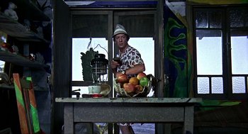 Movie still from “Age of Consent” (1969), directed by Michael Powell – A man standing in front of a table holding a basket of fruit; Wide shot, Over the shoulder angle