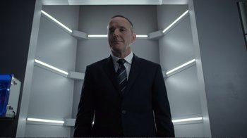 Movie still from “Agents of S.H.I.E.L.D.” (2013), created by Joss Whedon – A man wearing a suit and tie standing in a room; Medium shot, Low angle