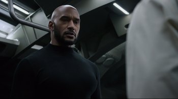 Movie still from “Agents of S.H.I.E.L.D.” (2013), created by Joss Whedon – A bald man in a black shirt is looking at the camera; Medium shot, Low angle