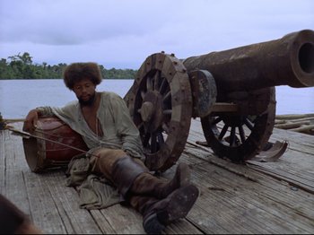 Movie still from “Aguirre, the Wrath of God” (1972), directed by Werner Herzog – A man sitting on a pier next to a cannon; Wide shot, Low angle