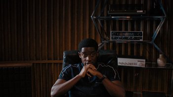 Movie still from “Snowfall” (2017), directed by Hiro Murai – A man sitting in a chair in front of a radio; Medium shot, High angle