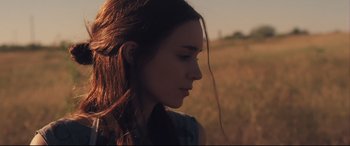 Movie still from “Ain't Them Bodies Saints” (2013), directed by David Lowery – A woman with long brown hair standing in a field; Close Up shot, Over the shoulder angle