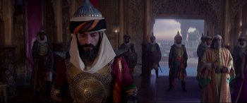 Movie still from “Aladdin” (2019), directed by Guy Ritchie – A man with a beard and a turban in a room with other men; Medium shot, Over the shoulder angle