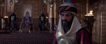Movie still from “Aladdin” (2019), directed by Guy Ritchie – A man in a costume stands in front of a staircase; Medium shot, Over the shoulder angle
