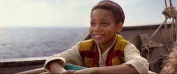 Movie still from “Aladdin” (2019), directed by Guy Ritchie – A young boy smiling for the camera while sitting on a pier; Close Up shot, High angle