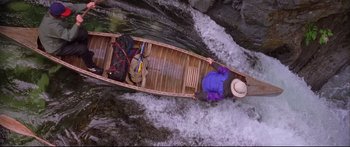Movie still from “Alaska” (1996), directed by Fraser C. Heston – A person in a boat with luggage on the side of a river; Extreme Wide shot, Overhead angle