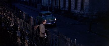 Movie still from “Alfie” (1966), directed by Lewis Gilbert – A man standing next to a car on a street; Wide shot, High angle