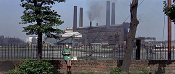 Movie still from “Alfie” (1966), directed by Lewis Gilbert – A woman sitting on a brick wall in front of a factory; Wide shot, Low angle