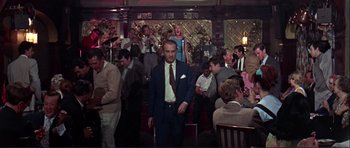 Movie still from “Alfie” (1966), directed by Lewis Gilbert – A man in a suit and tie standing in front of a group of people; Wide shot, Low angle