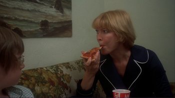 Movie still from “Alice Doesn't Live Here Anymore” (1974), directed by Martin Scorsese – A woman eating a slice of pizza while sitting on a couch; Medium shot, Over the shoulder angle