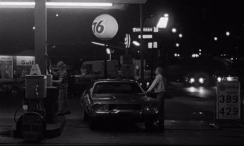 Movie still from “Alice in the Cities” (1974), directed by Wim Wenders – A man standing next to a car at a gas station at night; Wide shot, Over the shoulder angle