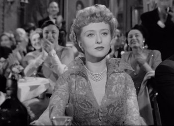 Movie still from “All About Eve” (1950), directed by Joseph L. Mankiewicz – An old black and white photo of a woman in front of a group of people; Medium shot, Low angle