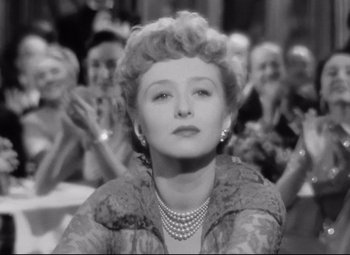 Movie still from “All About Eve” (1950), directed by Joseph L. Mankiewicz – An old black and white photo of a woman in front of a group of people; Close Up shot, Low angle