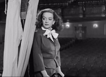 Movie still from “All About Eve” (1950), directed by Joseph L. Mankiewicz – A black and white photo of a woman standing in front of a curtain; Medium shot, Low angle