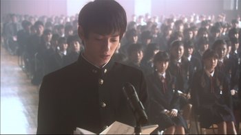 Movie still from “All About Lily Chou-Chou” (2001), directed by Shunji Iwai – A man in a black jacket is speaking to a crowd of people; Close Up shot, Over the shoulder angle