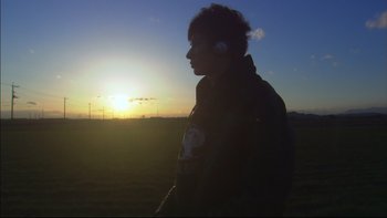 Movie still from “All About Lily Chou-Chou” (2001), directed by Shunji Iwai – A man with headphones standing in a field at sunset; Medium shot, Low angle