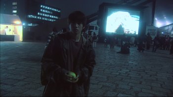 Movie still from “All About Lily Chou-Chou” (2001), directed by Shunji Iwai – A man holding an apple while standing in front of an outdoor screen; Medium shot, Over the shoulder angle