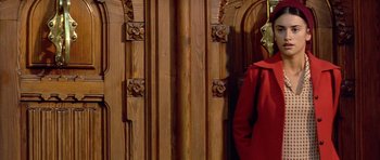 Movie still from “All About My Mother” (1999), directed by Pedro Almodóvar – A person wearing a red coat and a mask standing in front of a wooden door; Medium shot, Low angle