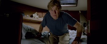 Movie still from “All Is Lost” (2013), directed by J.C. Chandor – An older man sitting on top of a bed holding a nintendo wii game controller; Medium shot, Low angle