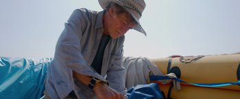 Movie still from “All Is Lost” (2013), directed by J.C. Chandor – An older man wearing a cowboy hat cutting a piece of fabric; Medium shot, Low angle