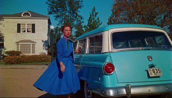 Movie still from “All That Heaven Allows” (1955), directed by Douglas Sirk – A woman in a blue dress standing in front of an old car; Wide shot, Low angle