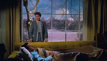 Movie still from “All That Heaven Allows” (1955), directed by Douglas Sirk – A woman standing next to a man laying on a couch; Wide shot, High angle