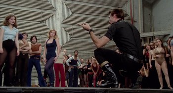 Movie still from “All That Jazz” (1979), directed by Bob Fosse – A man kneeling down on the ground in front of a group of people; Wide shot, Low angle