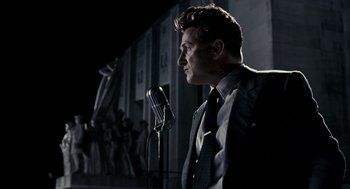 Movie still from “All the King's Men” (2006), directed by Steven Zaillian – A man standing in front of a microphone in a suit; Medium shot, Low angle