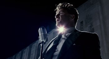 Movie still from “All the King's Men” (2006), directed by Steven Zaillian – A man in a suit and tie holding a microphone; Medium shot, Low angle