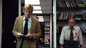 Movie still from “All the President's Men” (1976), directed by Alan J. Pakula – A man standing in an office holding a newspaper; Medium shot, Over the shoulder angle
