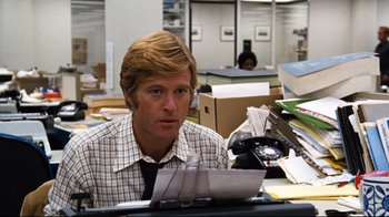 Movie still from “All the President's Men” (1976), directed by Alan J. Pakula – A man sitting at a desk in an office looking at a piece of paper; Medium shot, Over the shoulder angle