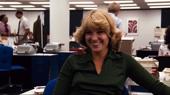 Movie still from “All the President's Men” (1976), directed by Alan J. Pakula – A woman sitting in an office chair smiling for the camera; Close Up shot, Over the shoulder angle