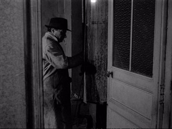 Movie still from “Alphaville” (1965), directed by Jean-Luc Godard – A man in a hat and overalls opening the door of a house; Wide shot, High angle