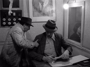 Movie still from “Alphaville” (1965), directed by Jean-Luc Godard – Two men are sitting at a table and one is smoking a cigarette and looking at a piece of paper; Medium shot, Over the shoulder angle
