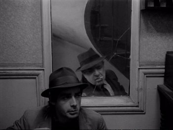 Movie still from “Alphaville” (1965), directed by Jean-Luc Godard – A black and white photo of a man in a hat looking in a mirror; Medium shot, Over the shoulder angle