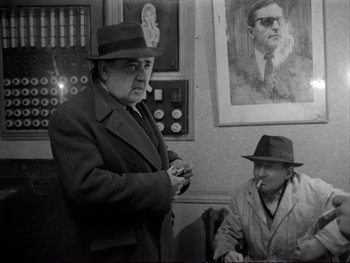 Movie still from “Alphaville” (1965), directed by Jean-Luc Godard – A man in a suit and hat next to another man in a hat; Medium shot, Over the shoulder angle