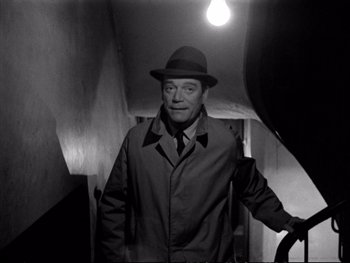 Movie still from “Alphaville” (1965), directed by Jean-Luc Godard – A black and white photo of a man in a fedora; Medium shot, High angle